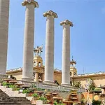 Čtyři vysoké sloupy, obklopené schodišti a zelení, stojí před architektonickou budovou na jasné modré obloze